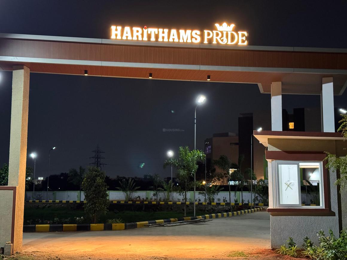 1000015575 The illuminated entrance gate of "HARITHAMS PRIDE" at night, featuring the prominent golden logo and name, a security cabin, and well-lit roads leading into the property.