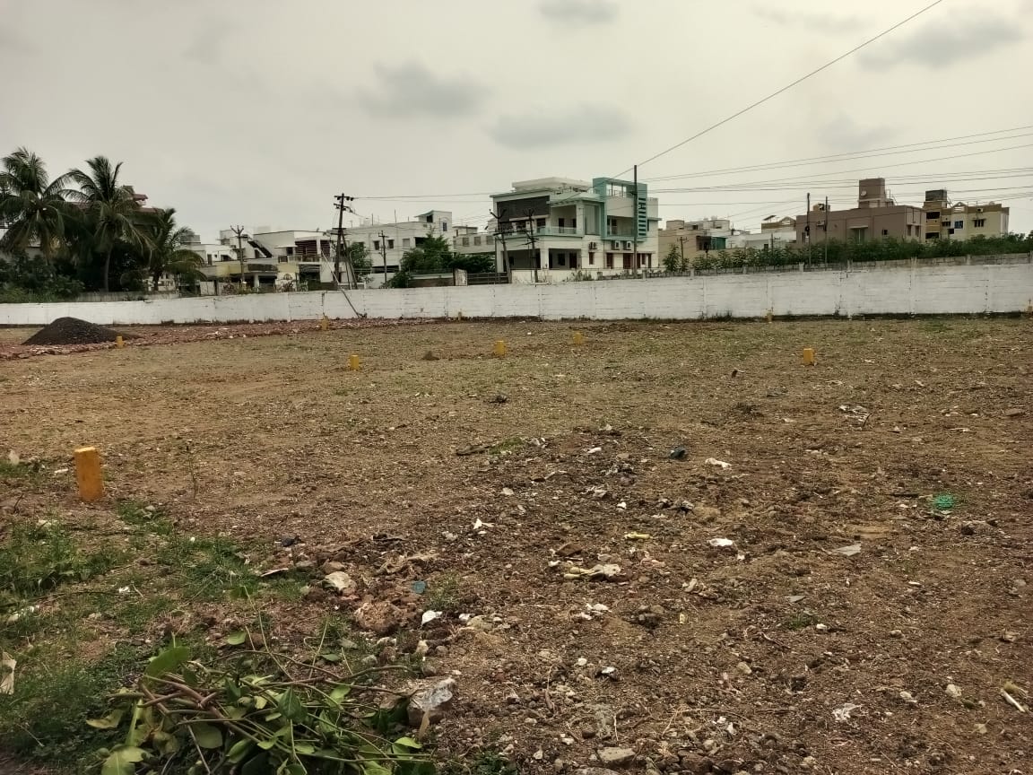 1000015613 A large, cleared residential plot marked with yellow boundary stones, bordered by a white wall and existing houses in the background, under an overcast sky.