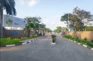 A wide-angle photo of the main entrance to a residential community called DivyaSree Hub6. The entrance features two large signboards and an avenue lined with trees and a central landscaped median. The signboard on the left reads "hub," "THE HUB OF ALL GOOD THINGS," "A community for 500+ families," and "Price starting at ~36 Lakhs." The bottom of the sign indicates "Hub 6 Urban Centre, OMR, Chennai." A contact number is also visible.