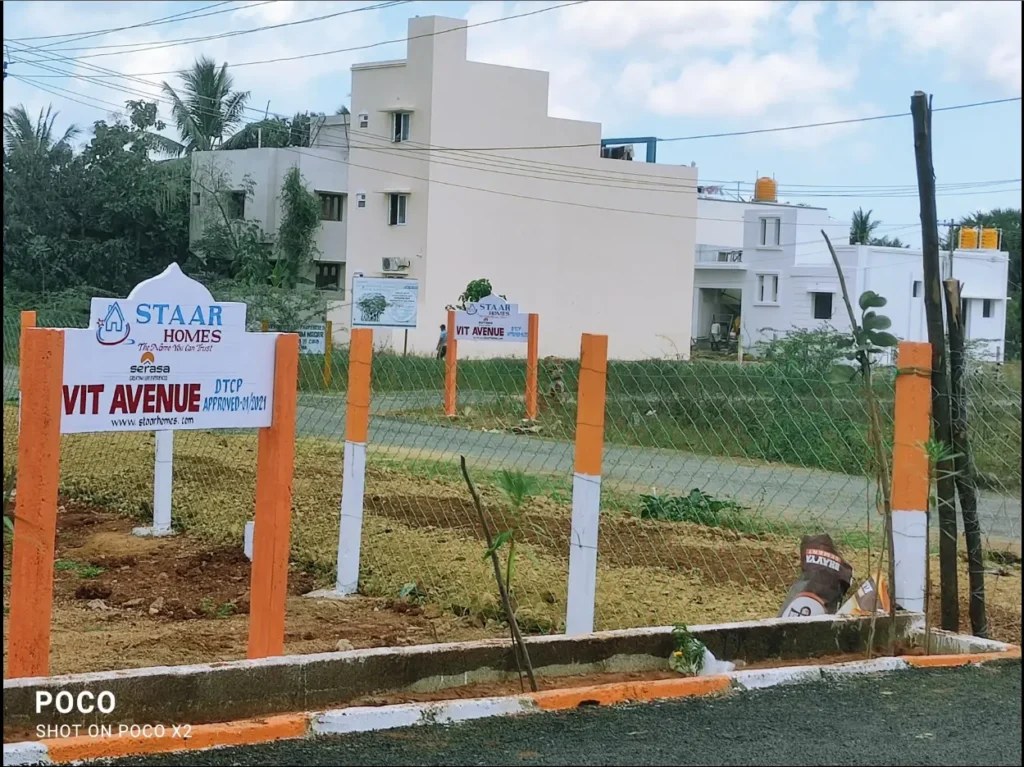 A photo of a residential land plot project called "Staar Homes VIT Avenue." A large, white and orange signboard in the foreground shows the project name, the Staar Homes logo, and the DTCP approval number (APPROVED-11/2021). The plot is marked by orange and white posts and a wire fence. In the background, there are a few residential buildings, a street, and a billboard with project details.