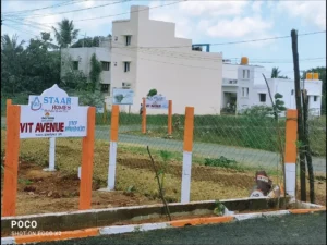 A photo of a residential land plot project called "Staar Homes VIT Avenue." A large, white and orange signboard in the foreground shows the project name, the Staar Homes logo, and the DTCP approval number (APPROVED-11/2021). The plot is marked by orange and white posts and a wire fence. In the background, there are a few residential buildings, a street, and a billboard with project details.