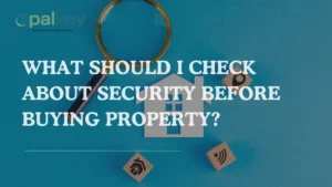 A graphic with a magnifying glass over a stylized paper house cutout, surrounded by three wooden blocks with security-related icons: a lock, an eye, and a wireless symbol. The main text asks, "WHAT SHOULD I CHECK ABOUT SECURITY BEFORE BUYING PROPERTY?". The background is a solid blue. The "opalkey" logo is in the top left corner.