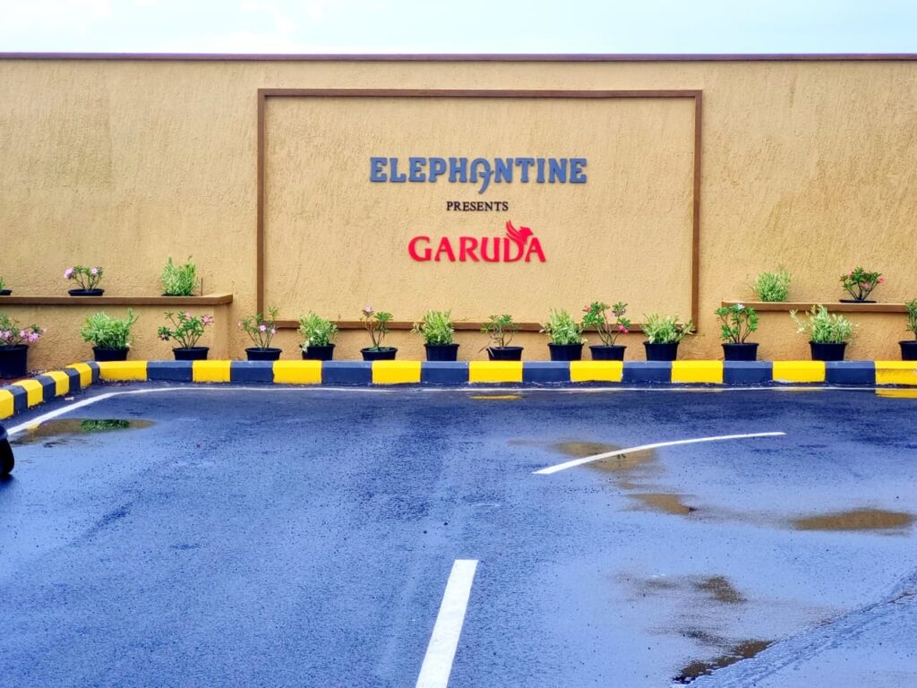 The main entrance wall of a residential project called "GARUDA" presented by "ELEPHANTINE." The name is displayed on a beige-colored textured wall, framed by a recessed border. Below the sign is a row of potted plants and a curb painted in yellow and black. The foreground is a wet, paved road with a white traffic line.