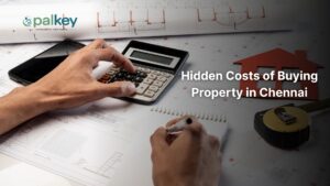 Hands using a calculator and notepad over property blueprints, next to a red model house and measuring tape. The text reads: "Hidden Costs of Buying Property in Chennai.