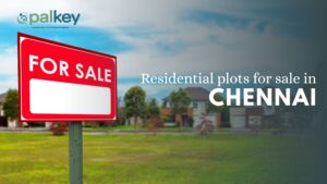 A promotional graphic for "Opalkey" with the text "Residential plots for sale in CHENNAI". The foreground features a large, red "FOR SALE" sign on a wooden post. The background is a blurry but lush green residential community with modern houses.